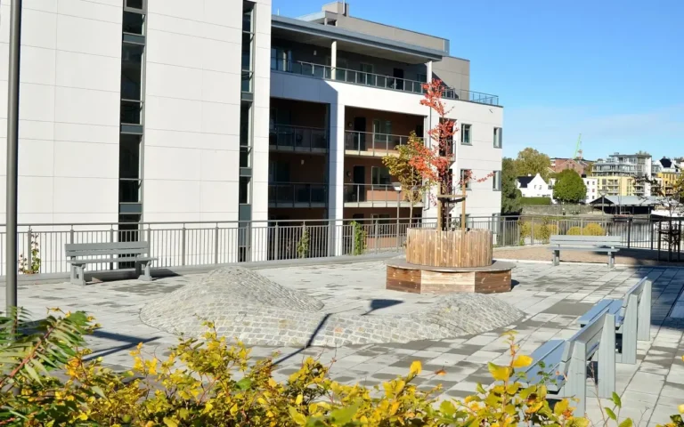 Mellom blokkene på Klosterøya ser du et område med steinbelegg og en rund benk med tre i midten.
